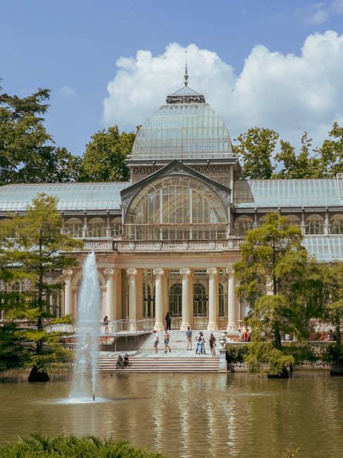 A large, historic greenhouse with a central glass dome and arched windows, situated on a landscaped property with tall trees and a pond in the foreground. There is a water fountain spraying upward in the pond, and people are visible walking on the steps and around the building, some carrying or standing near furniture and boxes. The scene is set outdoors on a partly cloudy day. The image reflects a setting where home relocation or packing activities could take place, with the building serving as a notable landmark in the background. The overall environment suggests a peaceful, scenic area suitable for residential or commercial moving services provided by Man and Van Crystal Palace, as indicated by the page context.