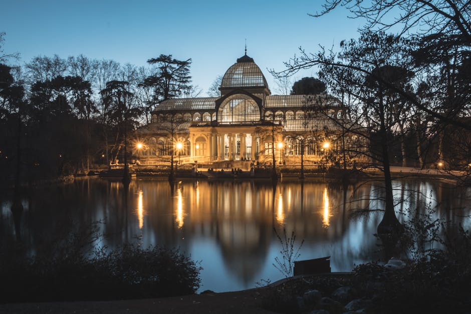 A large, ornate glass conservatory or greenhouse illuminated by exterior lighting at dusk, situated near a reflective body of water surrounded by trees and shrubbery. The structure features a domed roof, arched windows, and decorative architectural elements. The scene is part of a park or garden setting, with trees silhouetted against the evening sky. This environment suggests a peaceful, scenic location often associated with outdoor leisure or botanical displays. The lighting highlights the building’s glass and metal framework, creating reflections on the water's surface. In a house removals context, this image underscores the importance of careful planning when transporting delicate furniture or outdoor structures during a home relocation or furniture transport project, such as those offered by Man and Van Crystal Palace. The scene illustrates the peaceful surroundings that can be affected during moving logistics or packing and moving processes near such scenic landmarks.