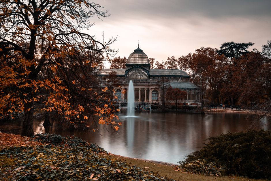 A large glass greenhouse with classical architectural features, including columns and a domed roof, situated next to a pond with calm water reflecting the structure. In the foreground, there are trees