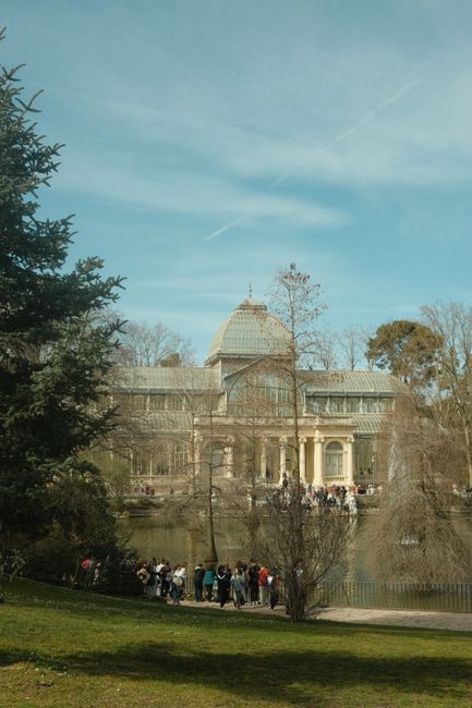 A large, historic greenhouse or conservatory with a glass dome roof and intricate architectural details, situated behind a park pond or small lake, with reflections visible in the water. In the foreground, a group of people are gathered along the water's edge, standing on a paved pathway. The scene is framed by trees and greenery, with some foliage directly in view, and the sky above is partly cloudy with patches of blue. The setting appears to be part of a botanical garden or conservatory complex in a park, with the building used for housing plants and supporting home relocation or moving activities nearby. The image captures the tranquil outdoor environment and the temporary gathering of visitors or workers near the historic building, consistent with a moving or packing process involving outdoor furniture or equipment.