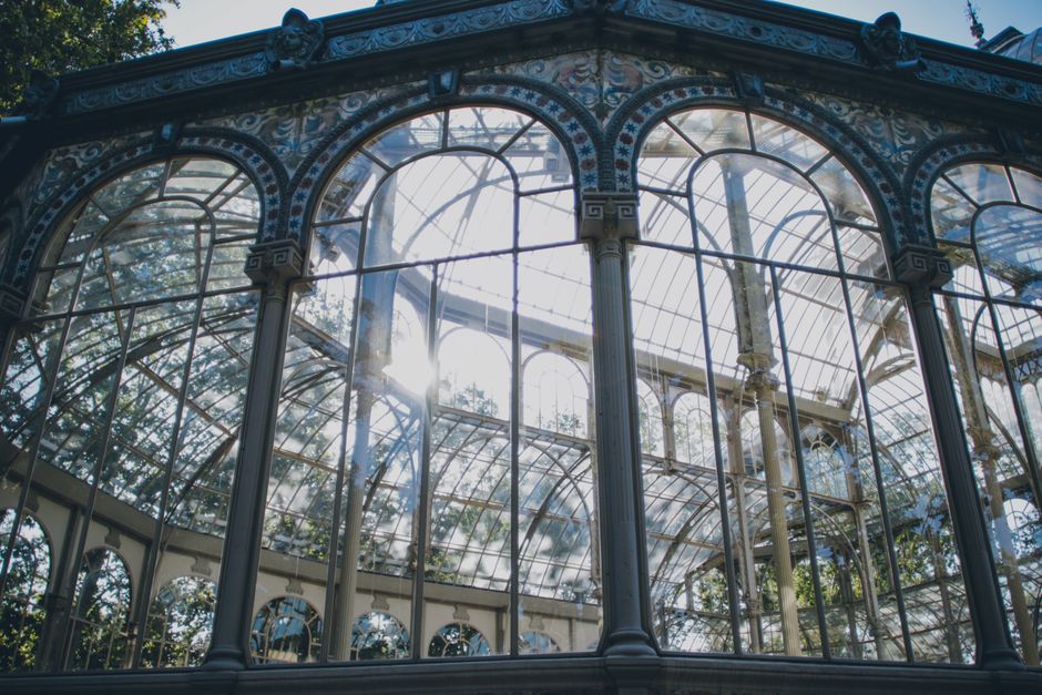 Photograph of an ornate glass conservatory structure with decorative metal framing and large arched windows, sunlight streaming through the glass panels. Inside and around the conservatory, there are