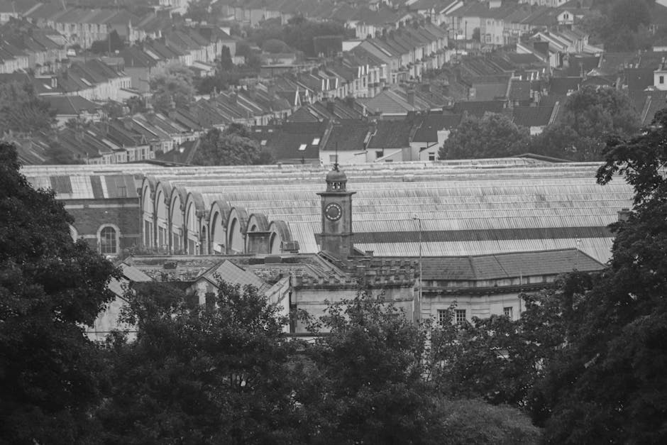 Photograph of an ornate glass conservatory structure with decorative metal framing and large arched windows, sunlight streaming through the glass panels. Inside and around the conservatory, there are